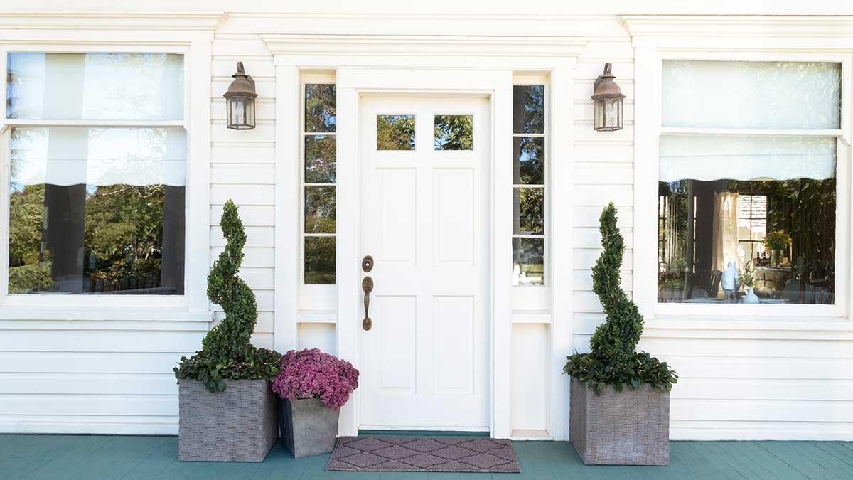 All white front door and windows with a pair of potted, spiraled Green Tower Boxwood plants and purple potted flowers.