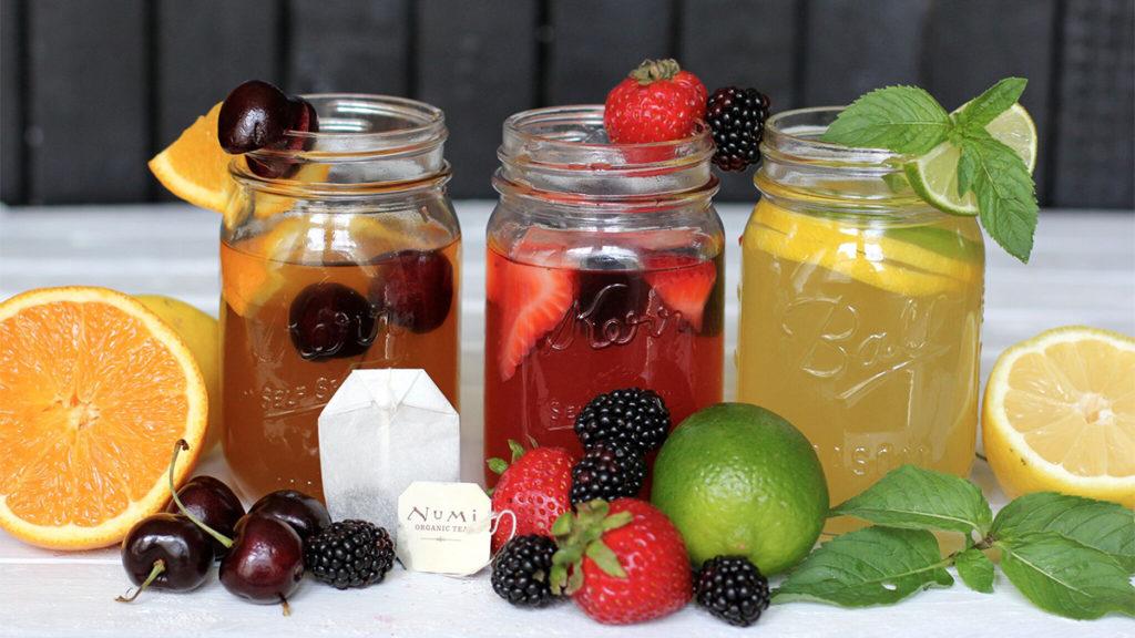 Three different Arnold Palmers in mason jars surrounded by fruit ingredients such as oranges, cherries, berries, and lemon.