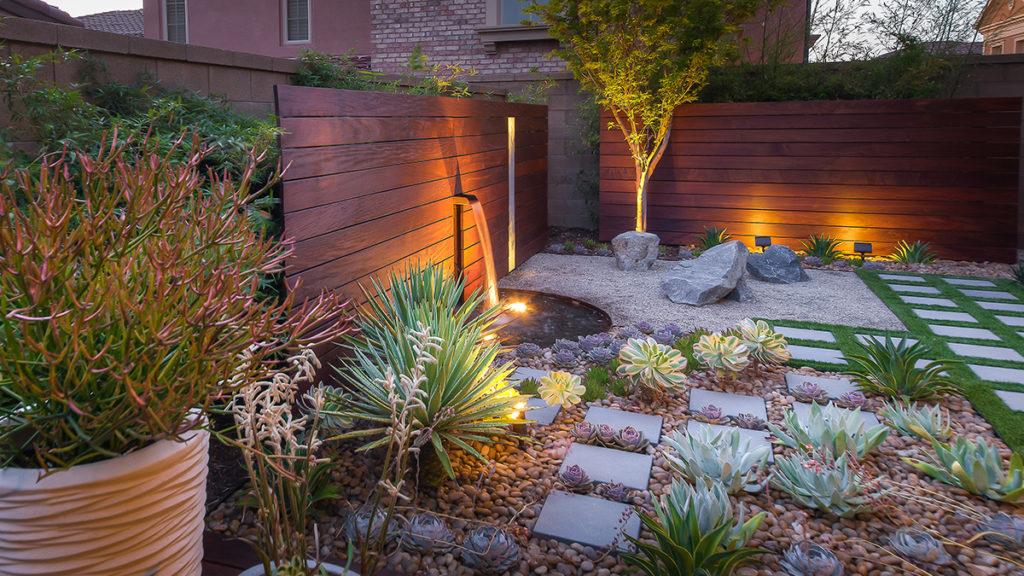 Tiny, water-wise backyard with a small fountain, potted plants, and succulents next to square stepping stones.