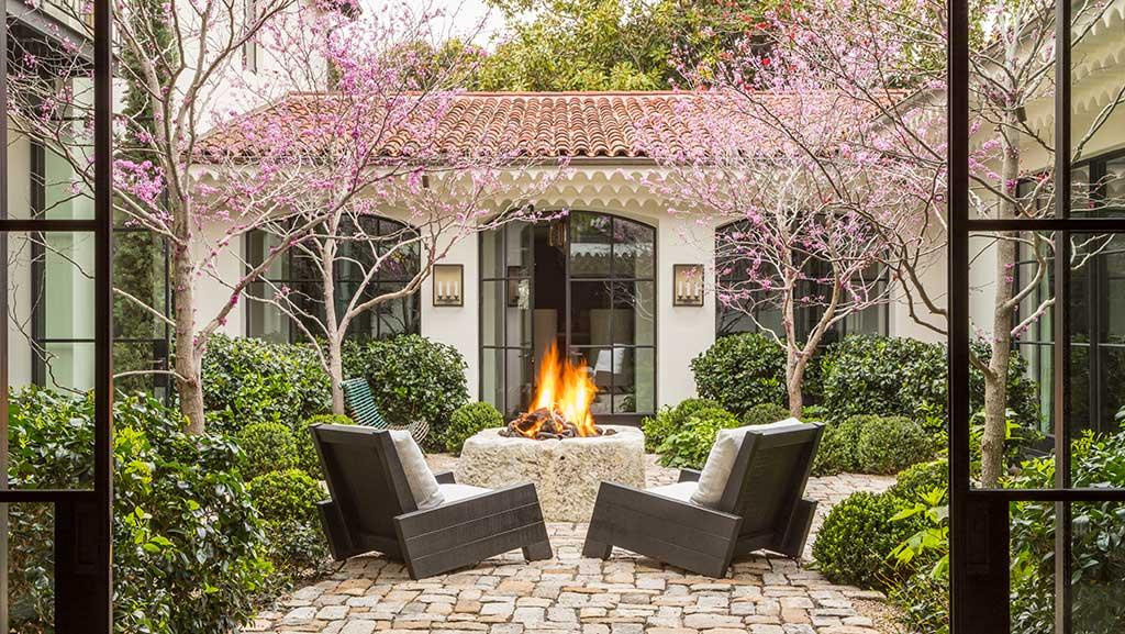 Courtyard with stone fireplace and two chairs in the center surrounded by sheared shrubs and trees with purple flowers. 