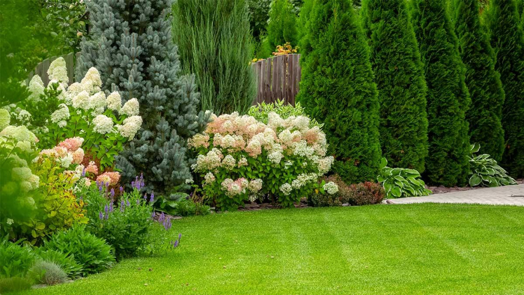 Green lawn with a border of tall trees and pink, white, and purple flowers including hydrangeas.