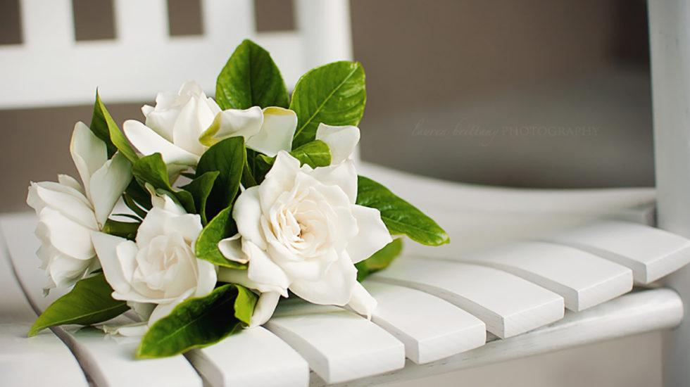 Arrangement of white gardenias and green leaves sitting on a white wooden chair.