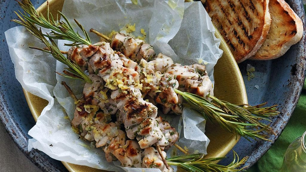 Chicken and rosemary skewers in a yellow dish plated next to two slices of bread on a blue plate.