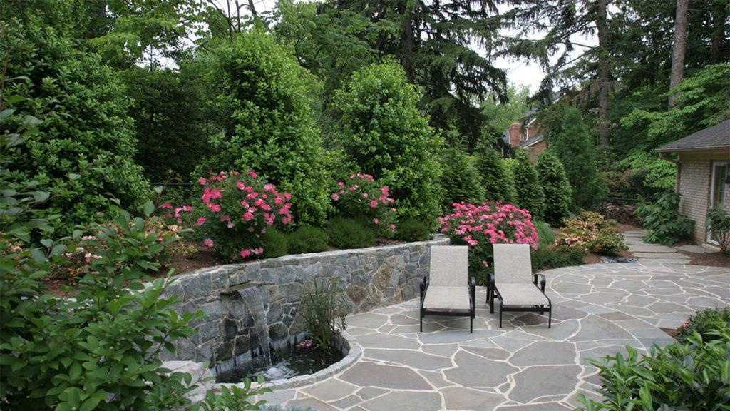 Backyard set near a forest with two lounge chairs surrounded by green trees, bushes, and pink flowers. 