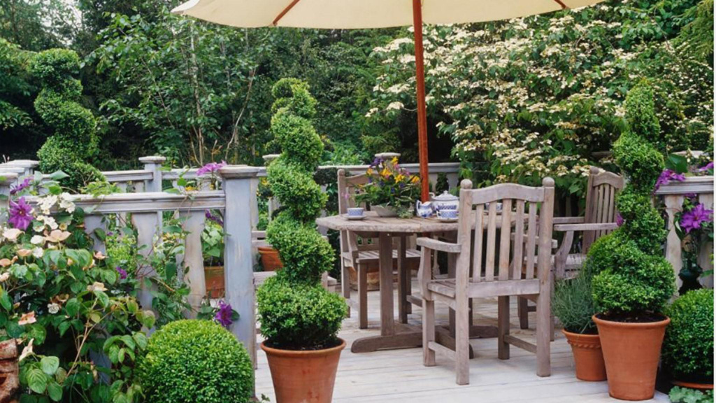 Patio with a wooden table and chairs surrounded by spiral topiaries in pots, trees, and purple and white flowers.