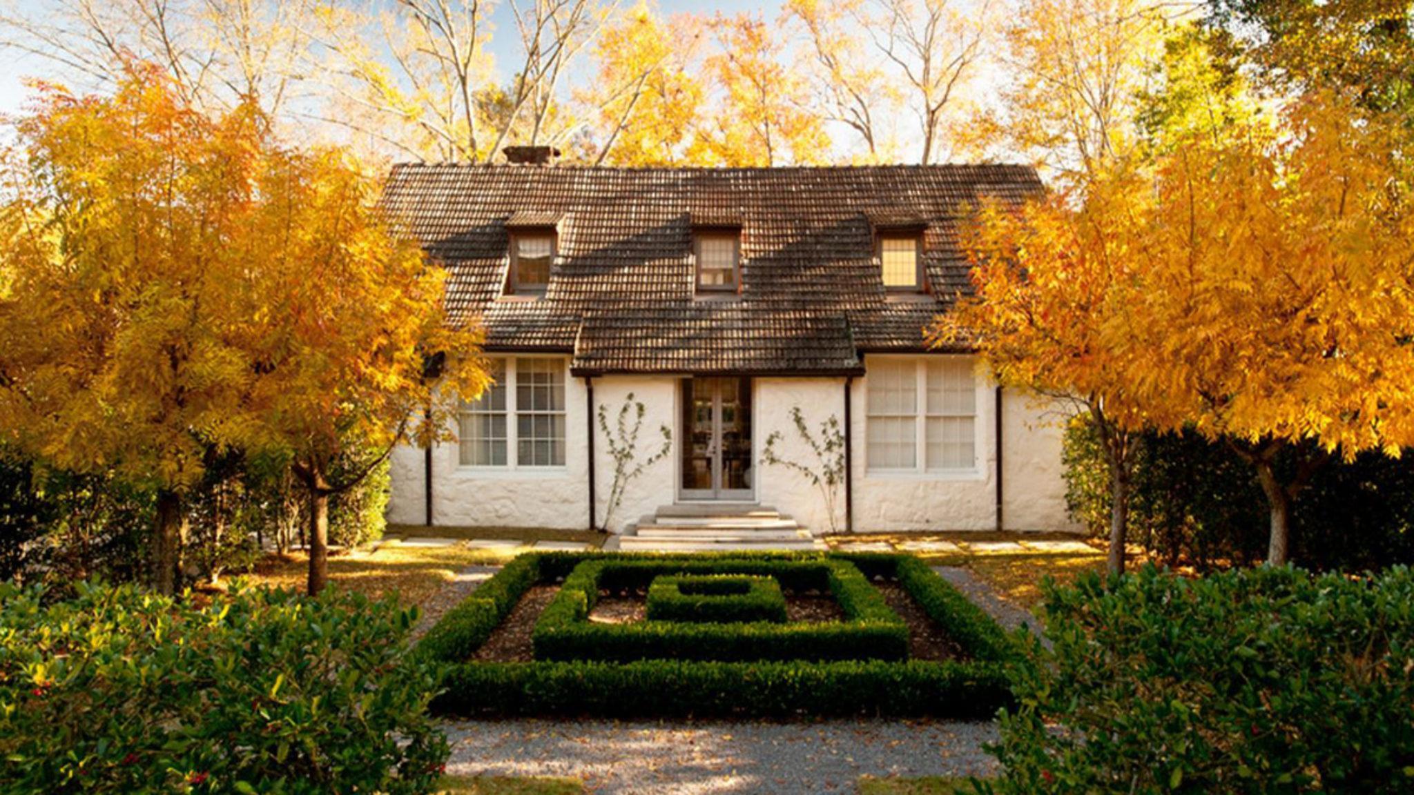 White cottage home with a garden full of golden-leafed trees and a maze style boxwood shrub in the center.