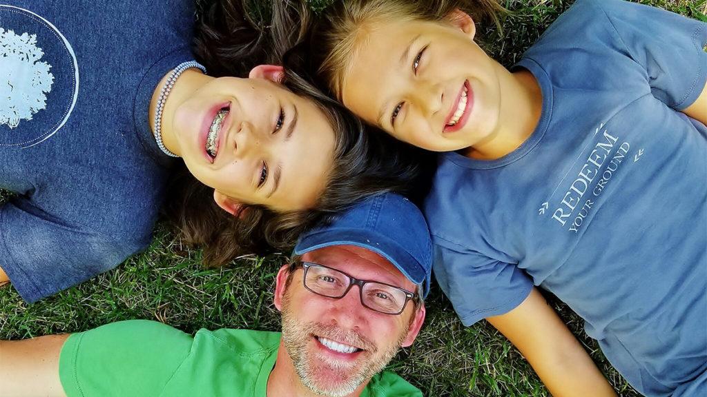 Birds eye view of a father with two young daughters smiling at the camera as they lay in the grass in a circle. 