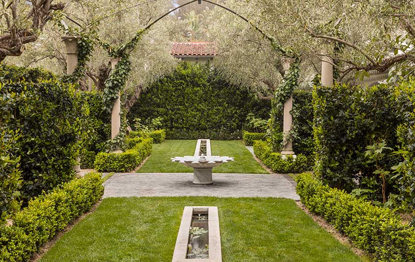 Symmetrical courtyard landscape featuring a green lawn, small shurbs, stephanotis vines, and a stone fountain.